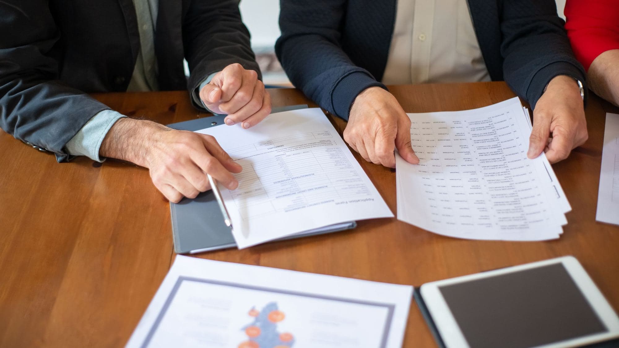 Hands on a wooden meeting table: one person points a pen at a line on a printed sheet, another holds a stack of printed pages, with a tablet and a printed chart in the foreground.