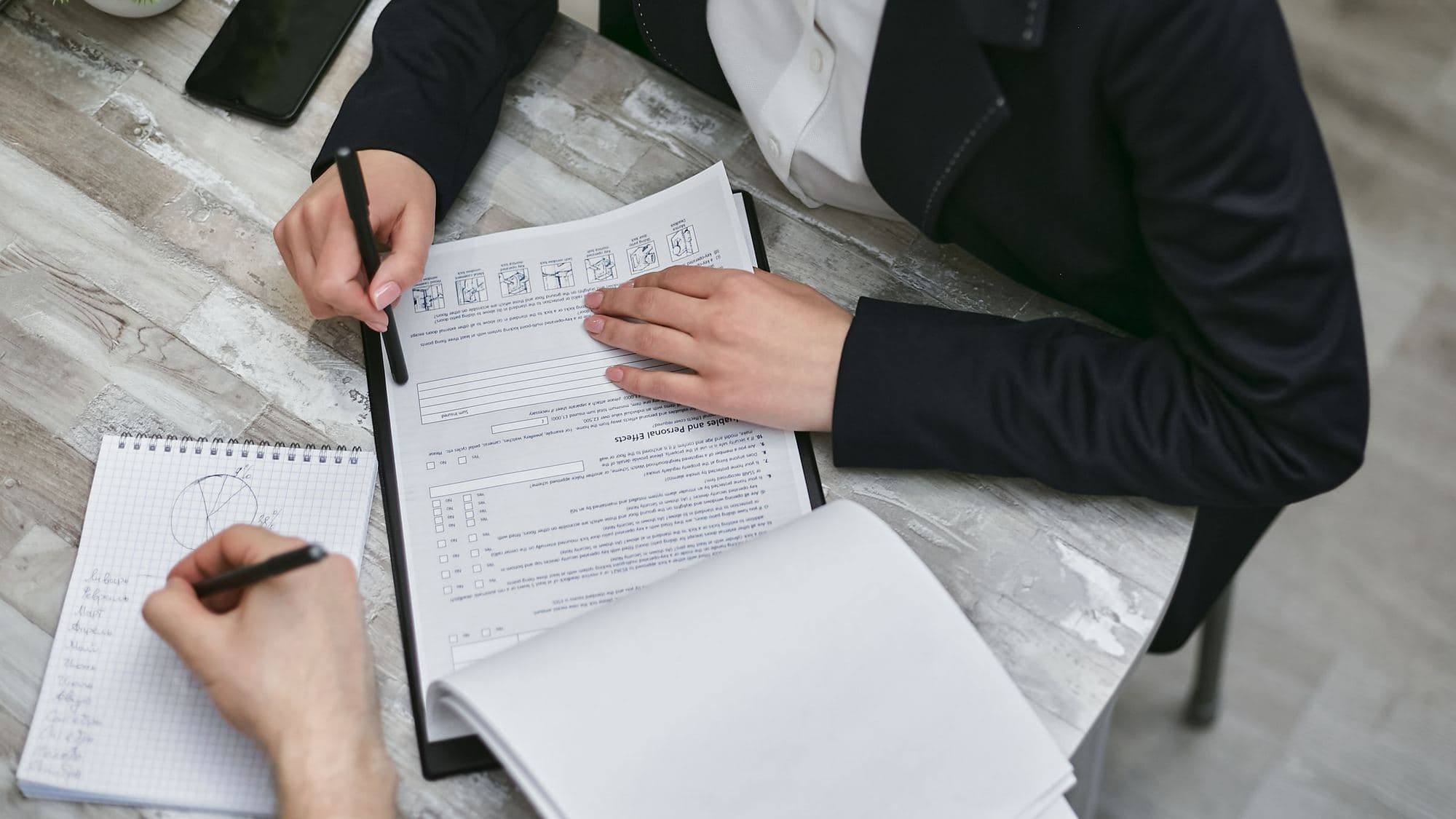 Seen from above, two people at a pale wooden desk: one in a dark suit holds a pen over a printed form on a clipboard, while the other writes in a spiral notebook.