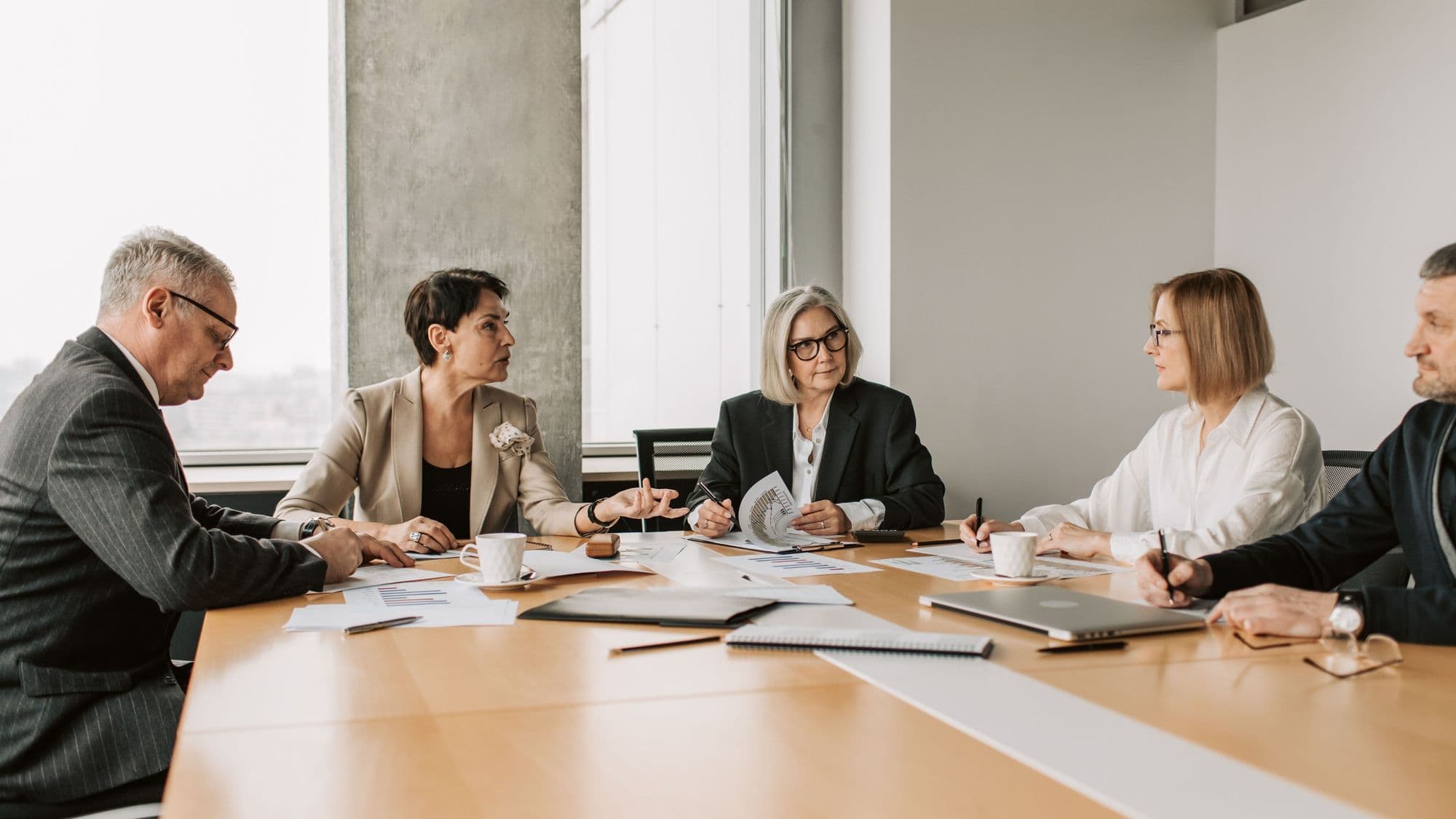 Five board members around a pale wooden table in a bright meeting room. One woman speaks and gestures while the others listen, two of them with pens over their papers.