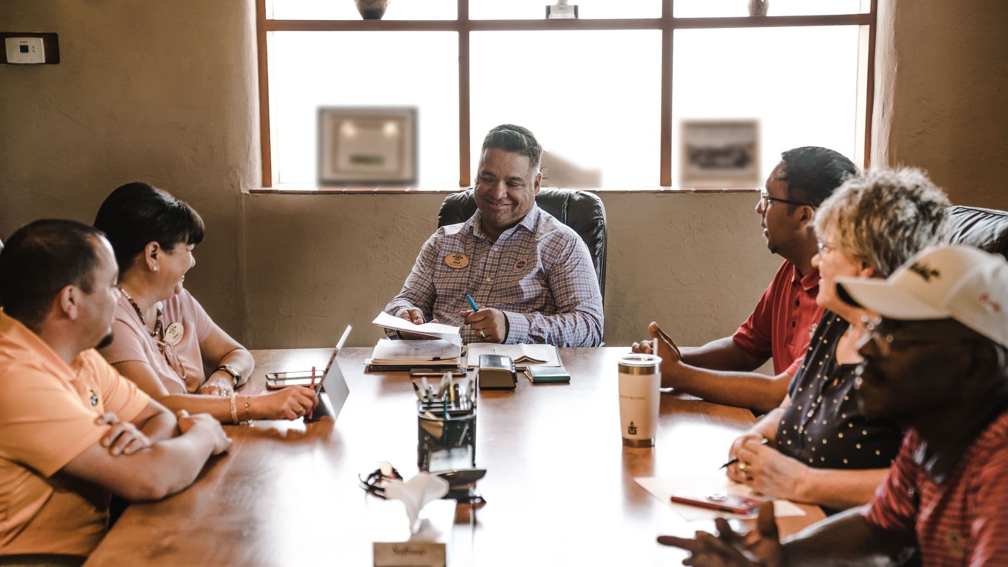 About seven people around a long wooden table in a plain, sunlit meeting room. The chair, at the head of the table, smiles as he passes a sheet of paper across.