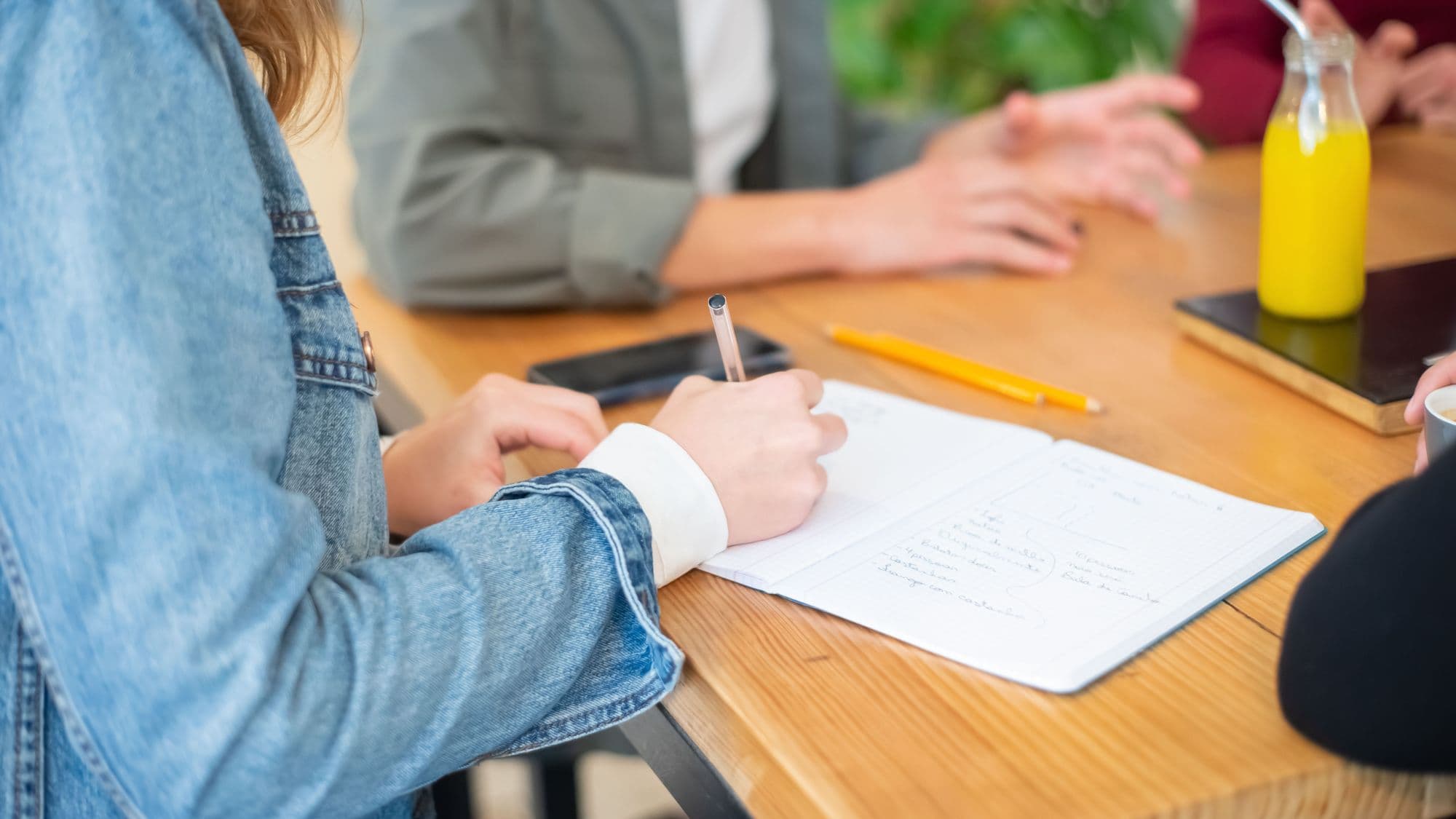 Over the shoulder of someone in a denim jacket writing in an open notebook at a wooden table, while two people talk across it.
