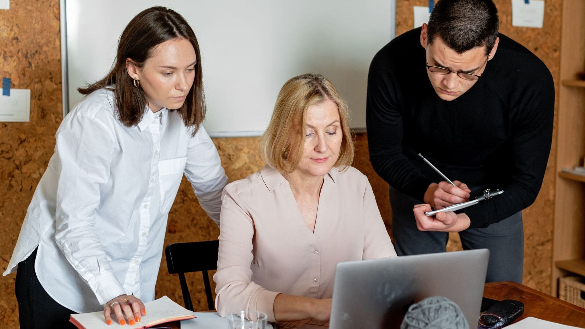 Three colleagues around a laptop in an office with a corkboard wall: a woman seated at the screen, another standing beside her with an open notebook, and a man in glasses writing on a clipboard.