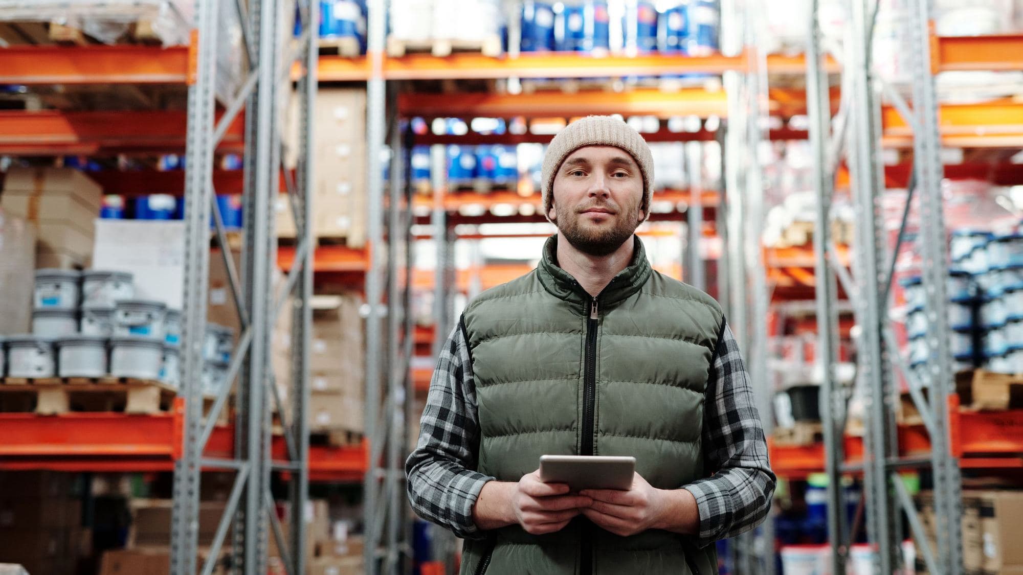 A warehouse worker in a beanie and padded gilet holds a tablet, standing in an aisle between tall orange racking stacked with drums and boxes.