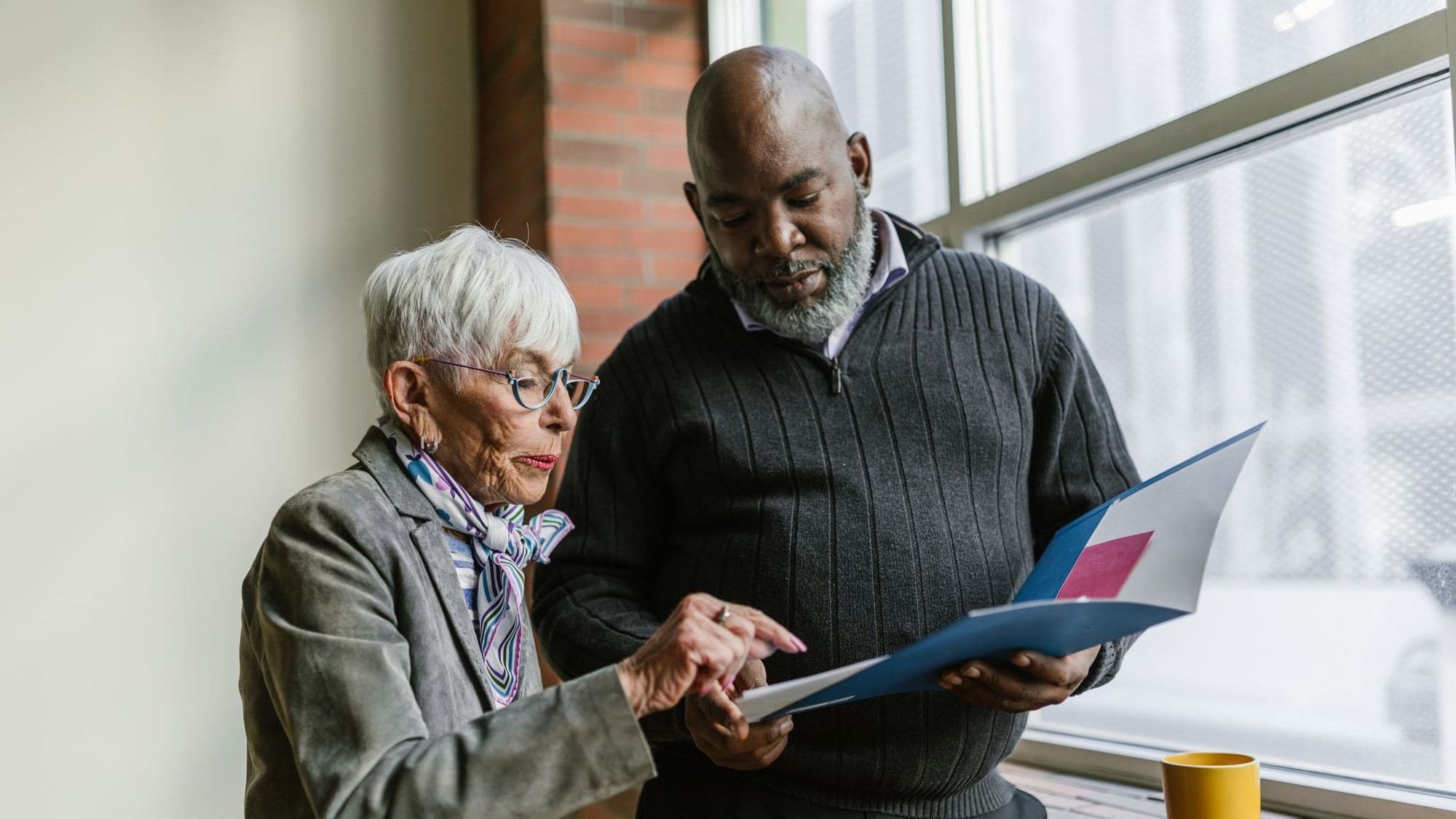 An older woman in a grey blazer and silk scarf points to a page in an open blue folder held by an older man in a dark sweater, as they stand reading it together by a tall window.